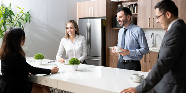Businesspeople standing and sitting around a table in an office break room with coffee.